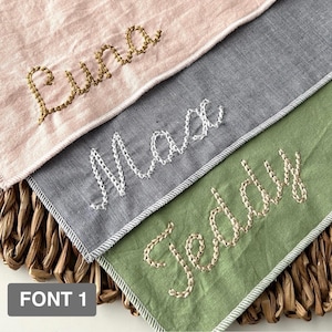 May include: Close-up of three personalized fabric items with embroidered names: "Luna" in gold, "Max" in white, and "Teddy" in beige. Each item has a contrasting border and is set against a woven background. The text "FONT 1" is at the bottom.