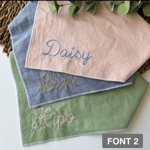 May include: Three personalized pet bandanas in pink, blue, and green, each embroidered with a name in a cursive font. The pink bandana has "Daisy" embroidered in blue thread. The blue bandana has "Blue" embroidered in gold thread. The green bandana has "Kipo" embroidered in pink thread.