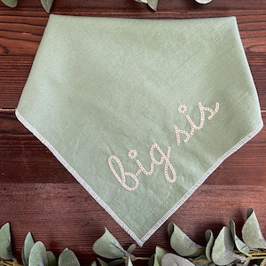 May include: A sage green bandana with the embroidered text "big sis" in a cursive font. The bandana has a white stitched border and is made of a soft fabric. The bandana is a baby accessory.