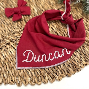 May include: A red dog bandana with the name "Duncan" embroidered in white. A matching red bow tie is also visible. The bandana is knotted at the top and has a white stitched border. The items are on a woven surface.
