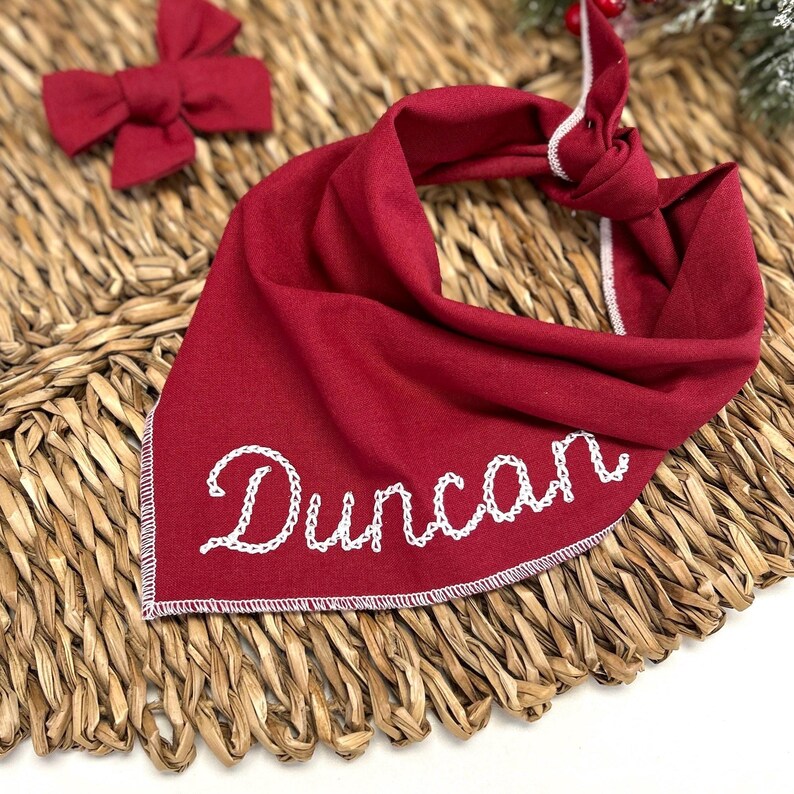 May include: A red dog bandana with the name "Duncan" embroidered in white. A matching red bow tie is also visible. The bandana is knotted at the top and has a white stitched border. The items are on a woven surface.