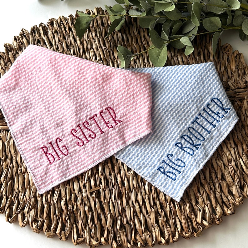 May include: Two seersucker bandanas, one pink and one blue, embroidered with "BIG SISTER" and "BIG BROTHER" in coordinating thread. The bandanas are placed on a woven, brown surface with green foliage in the background.