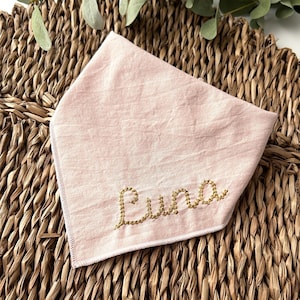May include: A light pink dog bandana with the name "Luna" embroidered in gold thread. The bandana is made of soft fabric and is folded in a triangle shape. The background is a woven wicker mat.