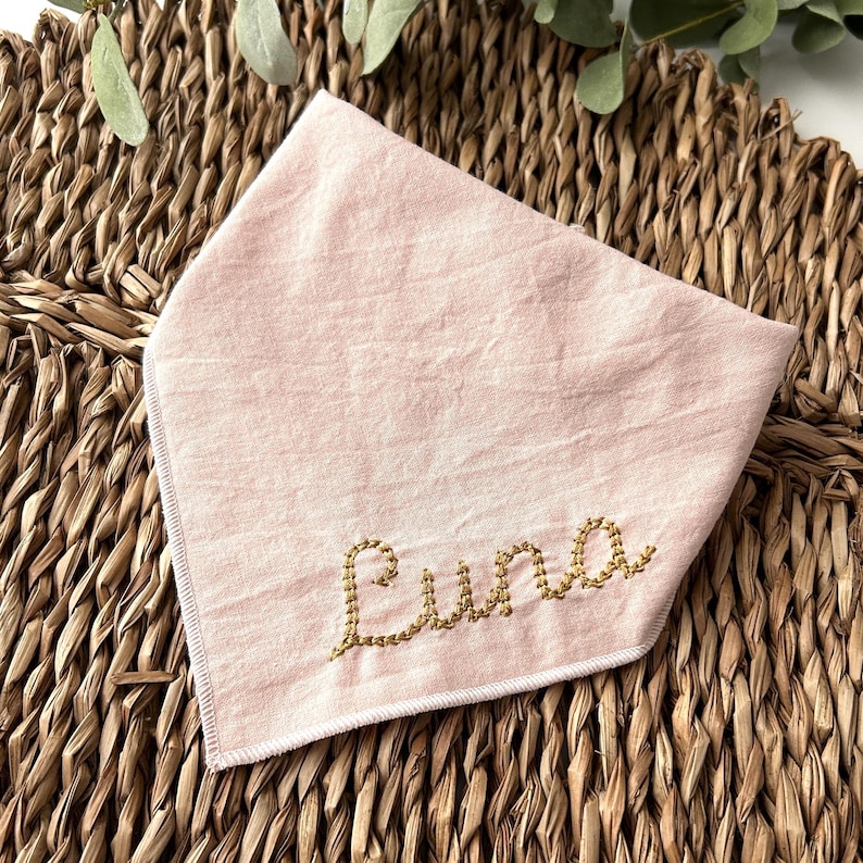 May include: A light pink dog bandana with the name "Luna" embroidered in gold thread. The bandana is made of soft fabric and is folded in a triangle shape. The background is a woven wicker mat.