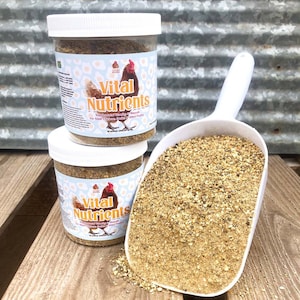K&ouml;nnte beinhalten: Zwei wei&szlig;e Gl&auml;ser mit Vital Nutrients, einem Nahrungserg&auml;nzungsmittel mit Vitaminen und Mineralien f&uuml;r erwachsene H&uuml;hner, Enten und anderes Gefl&uuml;gel. Die Gl&auml;ser stehen auf einer Holzoberfl&auml;che mit einer wei&szlig;en Schaufel voller Produkt.