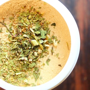 Puede incluir: Un taz&oacute;n blanco lleno de un polvo marr&oacute;n claro y hierbas verdes. Las hierbas son una mezcla de hojas y tallos secos.