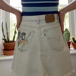 May include: A pair of light blue denim shorts with a white embroidered design of a face with glasses and flowers on the back pocket.