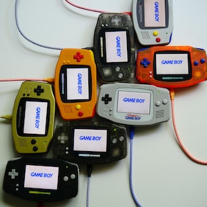 Könnte beinhalten: Eine Sammlung von Game Boy-Konsolen in verschiedenen Farben, darunter Orange, Grün, Schwarz und Grau. Jede Konsole zeigt "GAME BOY" auf dem Bildschirm an. Kabel in verschiedenen Farben verbinden die Konsolen.