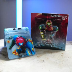 Op de afbeelding: Een lichtblauwe Game Boy Advance SP-console met een Metroid-afbeelding op de voorkant, naast een bijpassende doos. De doos bevat de tekst "Game Boy Advance SP" en een Metroid-karakterillustratie. Het Nintendo-logo is zichtbaar.