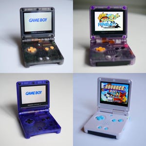 May include: Four Game Boy Advance SP consoles in different colors: translucent gray, purple, blue, and white. Each console is open, displaying a game on the screen. The screens show the words "GAME BOY" or a game title.