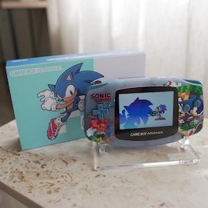 May include: A Game Boy Advance handheld console with a blue and white Sonic the Hedgehog design. The console is displayed on a clear stand next to its matching box. The box features a large image of Sonic.