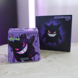 May include: A purple Game Boy Advance SP console with a Gengar design. The console features a Gengar graphic with a white grin and pink eyes. The box behind the console has a Gengar graphic and the text "GAME BOY ADVANCE SP".