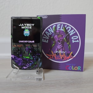 Puede incluir: Una consola de juegos portátil Game Boy Color negra y morada con un diseño personalizado que presenta al personaje Unit-01 de la serie de anime Neon Genesis Evangelion. La consola tiene un soporte de plástico transparente y una pantalla que muestra el texto "JAYBOY MODZ" y "GAME BOY COLOR".