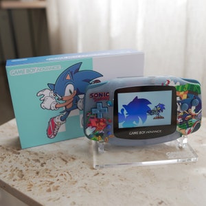 May include: A blue Game Boy Advance handheld console with a Sonic the Hedgehog design. The console is on a clear plastic stand and has a screen showing a blue Sonic character. The box for the console is in the background and features a blue Sonic character running.