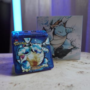 Op de afbeelding: Een blauwe, transparante Pokémon Game Boy-hoes met een Blastoise-afbeelding. De hoes heeft de Pokémon- en Nintendo-logo's. Een bijpassende doos met een Blastoise-illustratie staat achter de hoes.