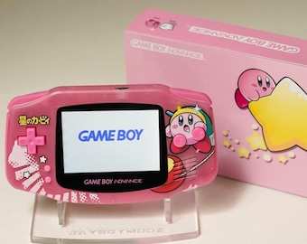 Puede incluir: Una consola portátil Game Boy Advance rosa con un diseño personalizado de Kirby. La pantalla muestra "GAME BOY" en azul. Una caja rosa a juego está detrás de la consola. La consola está sobre un soporte transparente.
