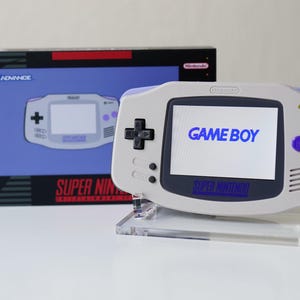May include: A Game Boy Advance handheld console in a light gray and purple color scheme, with the words "GAME BOY" displayed on the screen. The console is next to its box, which features the words "GAME BOY ADVANCE" and "SUPER NINTENDO".