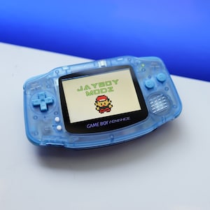 May include: A translucent blue Game Boy Advance handheld gaming console with a black screen displaying the text "JAYBOY MODZ" and a pixelated image of a video game character. The console has a purple "GAME BOY ADVANCE" logo.