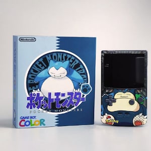May include: A Game Boy Color console and box set featuring a Snorlax design. The box is light blue and dark blue with Japanese text and the Nintendo logo. The console has a clear shell with a Snorlax graphic.