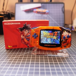 May include: An orange Game Boy Advance handheld console with a Dragon Ball Z theme, displayed on a clear stand. The screen shows the game's title. A red box with a matching design is visible to the left.