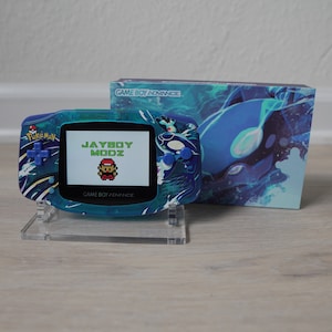 Puede incluir: Una Game Boy Advance SP azul con un diseño personalizado que presenta un patrón de olas azul y blanco. La pantalla muestra el texto "JAYBOY MODZ" y una imagen pixelada de un personaje de videojuego. La Game Boy Advance SP está en un soporte de acrílico transparente.