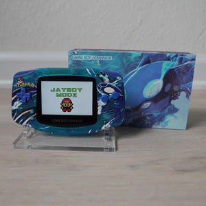 Könnte beinhalten: Eine türkisblaue Game Boy Advance-Konsole mit individuellem Pokémon-Design. Der Bildschirm zeigt "JAYBOY MODZ" und einen pixeligen Charakter. Die Konsole steht auf einem transparenten Ständer, mit einer passenden Box im Hintergrund.