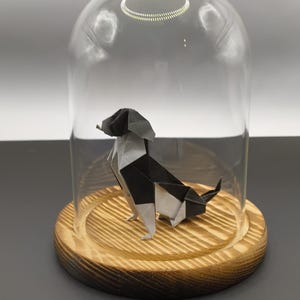 Può includere: Un cane origami bianco e nero sotto una campana di vetro. Il cane è posizionato su una base rotonda di legno, racchiusa da una cupola di vetro trasparente. La base in legno presenta venature naturali.