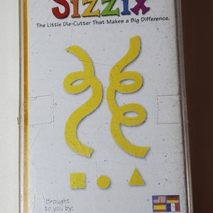 May include: A yellow die-cutting machine for crafting, with the brand name "Sizzix" and the text "The Little Die-Cutter That Makes a Big Difference." The die-cutting machine is designed to cut out confetti shapes, including a square, circle, and triangle. The die-cutting machine is brought to you by Provo Craft Ellison and has the product number 38-0148.