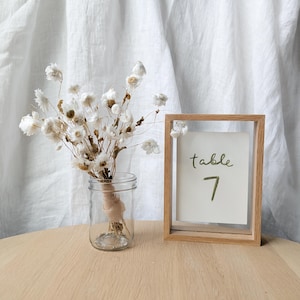 May include: A clear glass jar with dried white flowers tied with twine, and a wooden framed sign that reads "table 7" in green. The objects are on a light wood table, with a white curtain in the background.