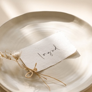 May include: A white plate with a small sprig of dried white flowers and a handwritten note that says "Ingrid" on a piece of white paper.