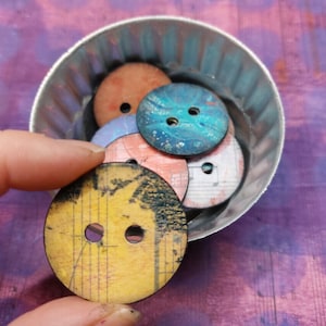 May include: A collection of colorful, patterned buttons in a silver metal container. The buttons are round and have two holes each. The buttons are made of wood and have a variety of designs, including musical notes, geometric patterns, and abstract art.