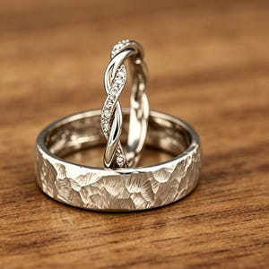Op de afbeelding: Een zilverkleurige ring met een brede, gehamerde textuurband ligt horizontaal, terwijl een dunnere, gedraaide zilverkleurige ring met kleine, heldere ingebedde stenen verticaal in het midden van de bredere band staat.