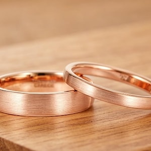 Könnte beinhalten: Zwei roségoldene Eheringe mit gebürsteter Oberfläche liegen auf einer Holzoberfläche. Die Ringe haben ein schlichtes, elegantes Design mit leicht abgerundetem Profil. Die Ringe liegen eng beieinander.