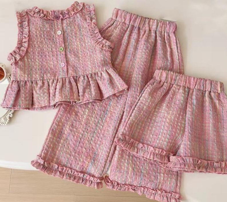 Puede incluir: Un conjunto de tres piezas de tweed rosa, que incluye una blusa sin mangas con volantes, botones y un peplum, adem&aacute;s de pantalones cortos y pantalones a juego. La tela tiene un sutil patr&oacute;n de hilo multicolor.