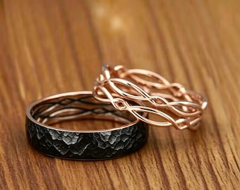 May include: A set of two rings consisting of one wide black band with a textured, hammered finish and one thinner rose gold band featuring an intricate, open-work braided or woven metal design that rests partially on the black ring.