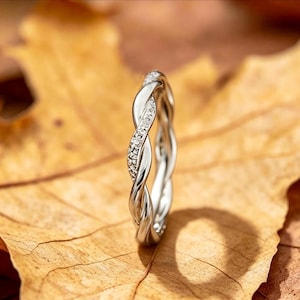 May include: A silver-coloured ring featuring a twisted, braided band design. One strand of the twisted metal is encrusted with small, clear, sparkling stones, while the other strand remains a smooth, polished metallic finish.