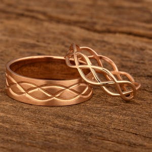 Puede incluir: Dos anillos de oro rosa. Un anillo tiene una banda ancha con un patrón de diamantes repetido. El otro anillo tiene un diseño abierto e entrelazado. Ambos anillos están sobre una superficie de madera.