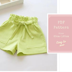 Puede incluir: Pantalones cortos verde lima con cintura alta fruncida y lazo a juego. Los pantalones cortos tienen dos bolsillos delanteros. Una tarjeta rosa a la derecha dice "PDF Pattern" con detalles de tallas y "Easy to sew".