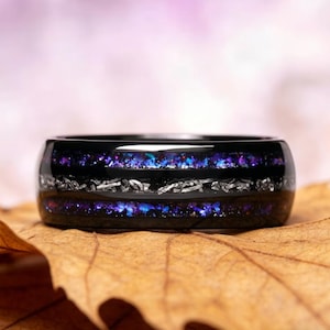 May include: A black ring with a silver band and a centre of purple and blue iridescent material. The ring is set on a brown leaf with a blurred pink background.