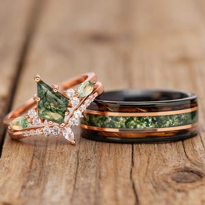 Può includere: Due anelli: un anello di fidanzamento in oro rosa con una gemma verde e piccoli diamanti, e una fascia nera con un intarsio verde e ramato. Gli anelli sono appoggiati su una superficie di legno.