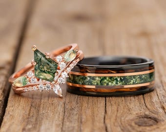 May include: Two rings: a rose gold engagement ring with a green gemstone and small diamonds, and a black band with a green and copper inlay. The rings are set on a wooden surface.