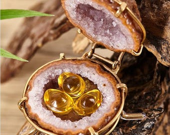 May include: A unique pendant featuring a geode design with a gold-colored metal frame. The pendant opens to reveal a cluster of three yellow, oval-shaped stones. The geode has a brown exterior and a purple crystal interior.