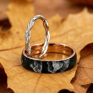 May include: A silver twisted band ring with small embedded stones resting against a wider ring featuring a black hammered exterior finish and a smooth rose gold interior band, showcasing a contrast between the textured and polished surfaces.