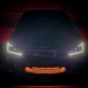 Może przedstawiać: Ciemny samochód z jasnymi reflektorami i świecącym pomarańczowym grillem. Przód samochodu jest wyśrodkowany w kadrze, z podświetlonymi reflektorami i świecącym grillem. Samochód wydaje się być w ciemnym otoczeniu.