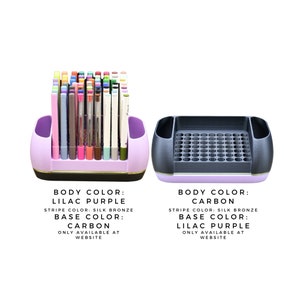 May include: Two pen holders, one lilac purple and one carbon. The lilac purple holder contains various pens. Both holders have a silk bronze stripe and a carbon or lilac purple base. Text on the image indicates availability on the website.