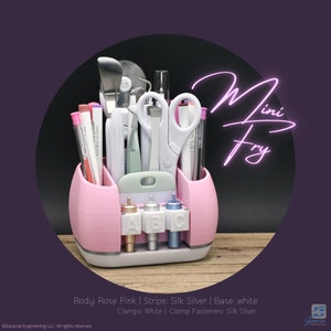 May include: A rose pink organizer with white and silver accents, holding various craft tools like scissors, pens, and a spatula. The organizer has the text "Mini Fry" in a neon style. The base is white, and the clamps are white with silver fasteners.