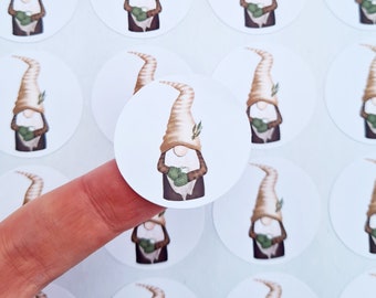 May include: A collection of round stickers featuring a whimsical gnome design. Each sticker showcases a gnome with a tall, pointed hat, holding a green bundle. The gnome wears a brown coat and has a white beard. The stickers are on a white background.
