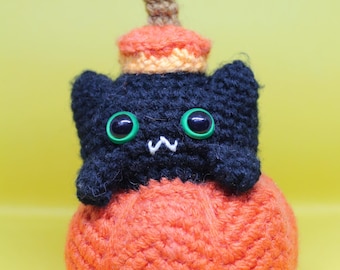 May include: A handmade crochet black cat with green eyes, sitting atop an orange pumpkin. The cat has a small white mouth and is wearing a pumpkin hat with a brown stem. The background is a solid yellow.