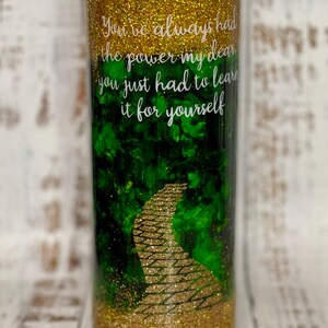 May include: A gold glitter tumbler with a green and gold glitter design of the yellow brick road. The tumbler has a white inscription that reads "You've always had the power my dear you just had to learn it for yourself."