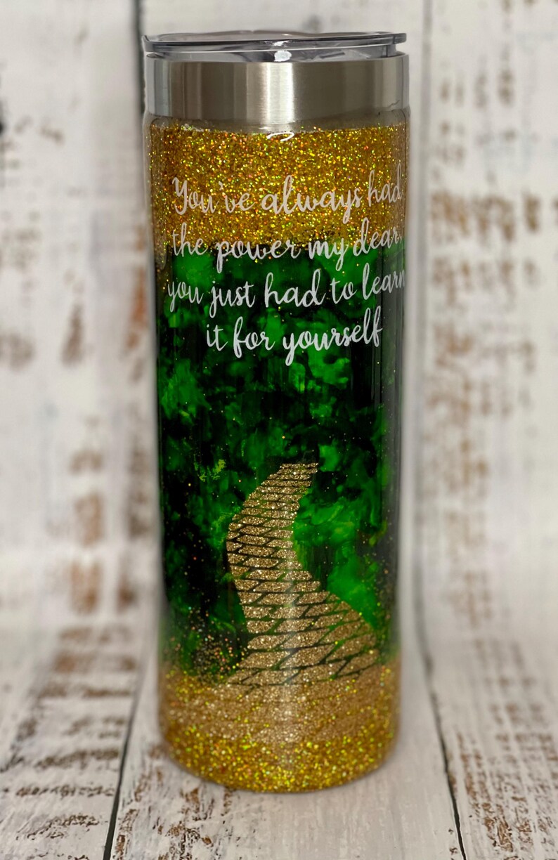 May include: A gold glitter tumbler with a green and gold glitter design of the yellow brick road. The tumbler has a white inscription that reads "You've always had the power my dear you just had to learn it for yourself."